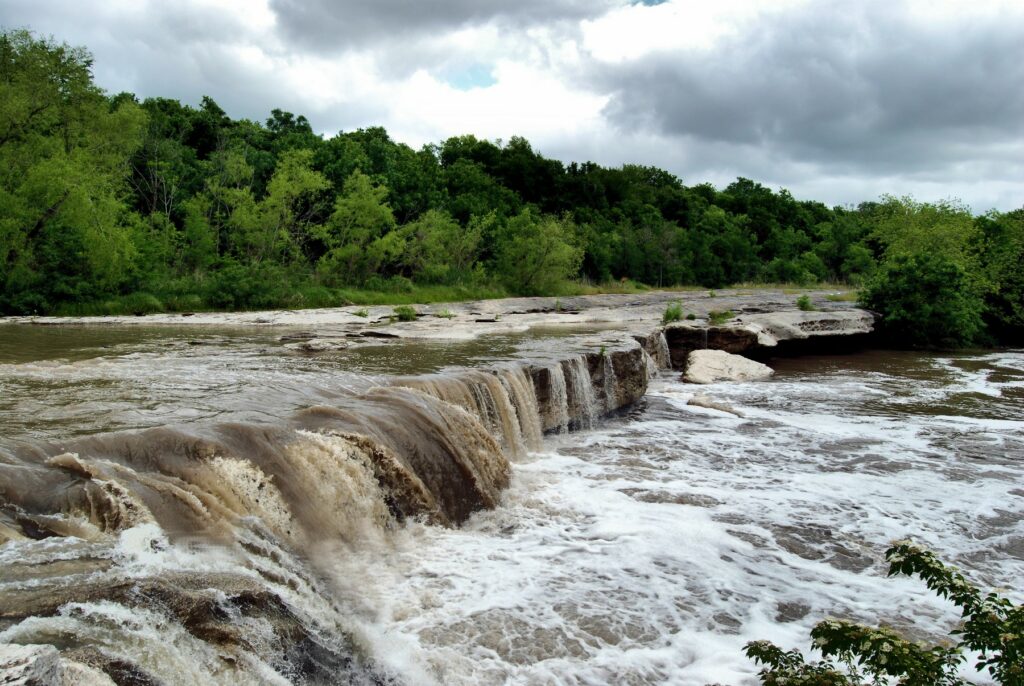 Waterfall at McKinney Falls State Park in Austin, Texas, showing a mix of turbulent water and lush greenery, highlighting the potential risks of flash flooding during extreme weather.