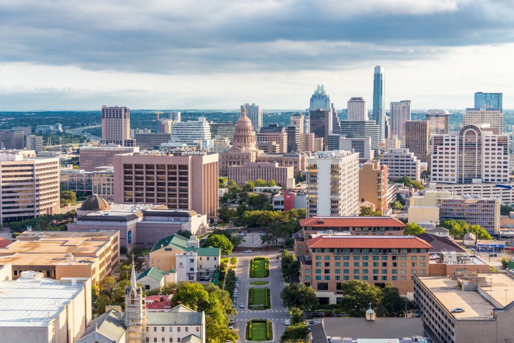 A panoramic view of downtown Austin, Texas, with iconic skyscrapers, the Texas State Capitol, and green spaces in the foreground, showcasing the city's vibrant urban landscape.