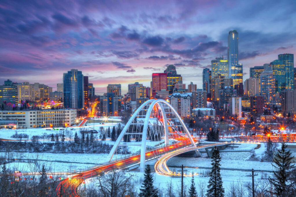 Light trails from rush hour traffic light up Edmonton downtown. Winter sunset skyline showing Walterdale Bridge across the frozen, snow-covered Saskatchewan River and surrounding skyscrapers.
