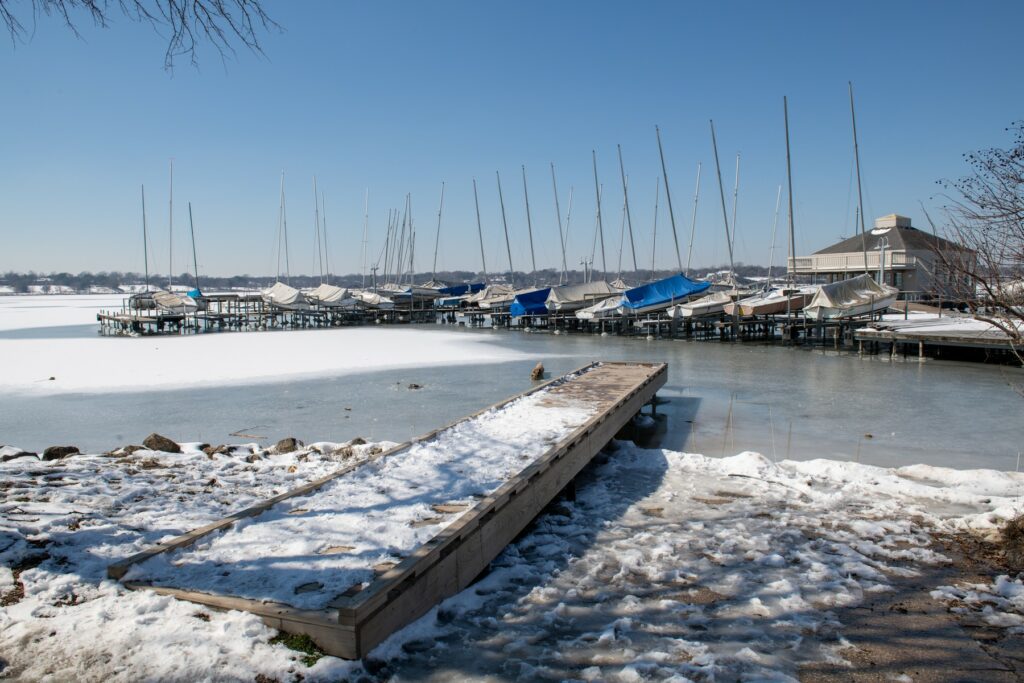 Snow-covered dock leading to a frozen lake, with sailboats docked along the shore and a clear winter sky, capturing the quiet beauty of Lakewood in the colder months.
