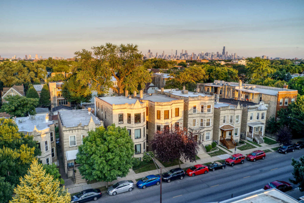 Aerial view of historic greystone homes in Logan Square, Chicago, with tree-lined streets and the city skyline in the background at sunset.