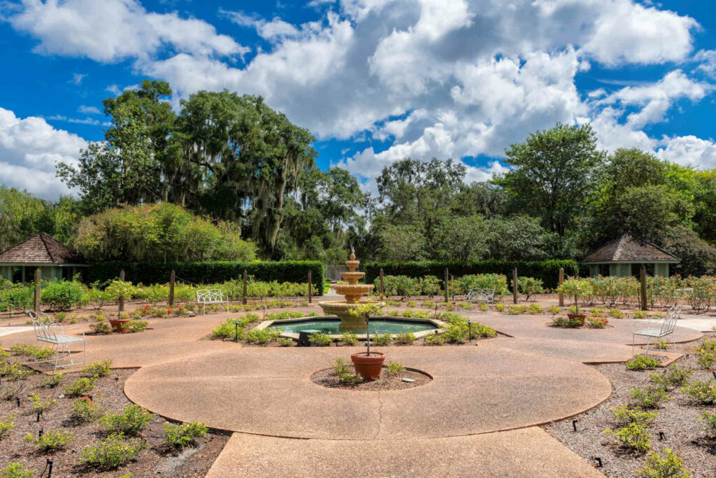 Peaceful garden with a central fountain, benches, and surrounding greenery in Meadow Woods, Florida, under a partly cloudy sky.