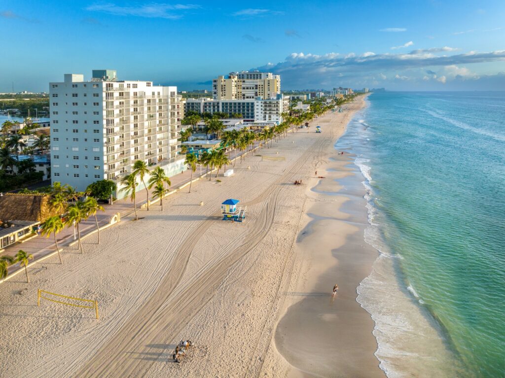 Aerial view of North Miami Beach with high-rise condos, palm-lined sand, and calm turquoise waters, showcasing a relaxed coastal lifestyle.