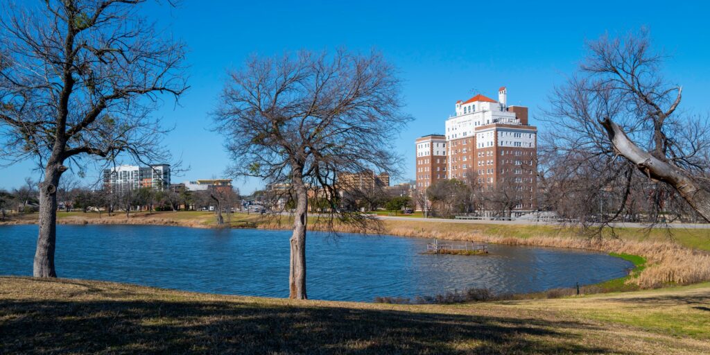 Scenic view of Oak Cliff with a tranquil lake, bare trees, and modern buildings in the distance, highlighting the neighborhood's mix of nature and urban development.