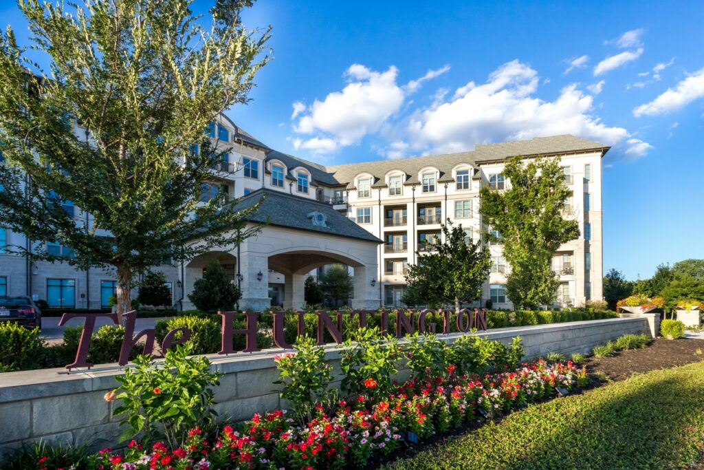 The Huntington apartment complex in Plano, Texas, with lush greenery, vibrant flowers, and a welcoming entrance, showcasing suburban living with modern amenities.