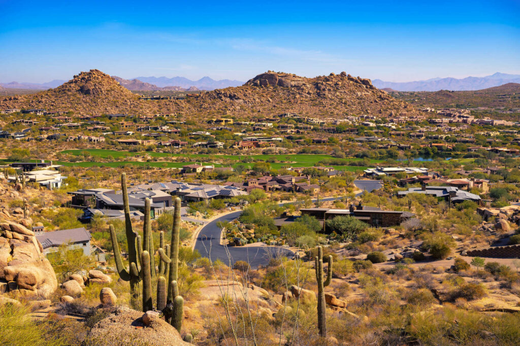 Scenic view of a suburban neighbourhood in Phoenix, Arizona, surrounded by desert landscape, mountains, and large saguaro cacti under a bright blue sky.