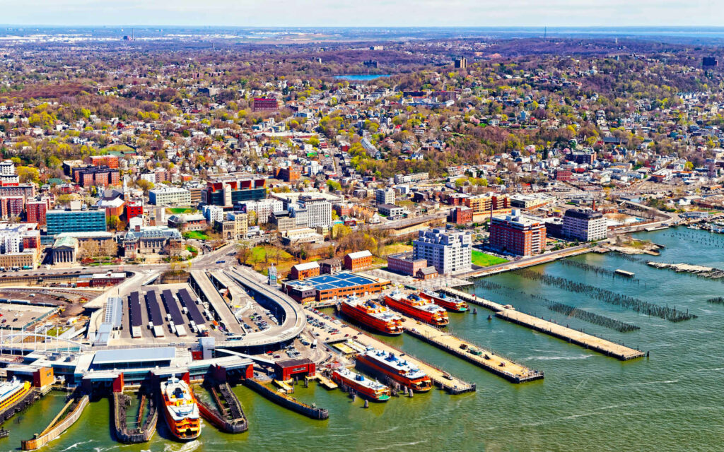 Aerial view of Staten Island's St. George Ferry Terminal, with orange ferries docked along the waterfront and a dense urban landscape in the background.